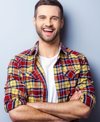 Happy young man. Portrait of handsome young man in casual shirt keeping arms crossed and smiling while standing against grey background
