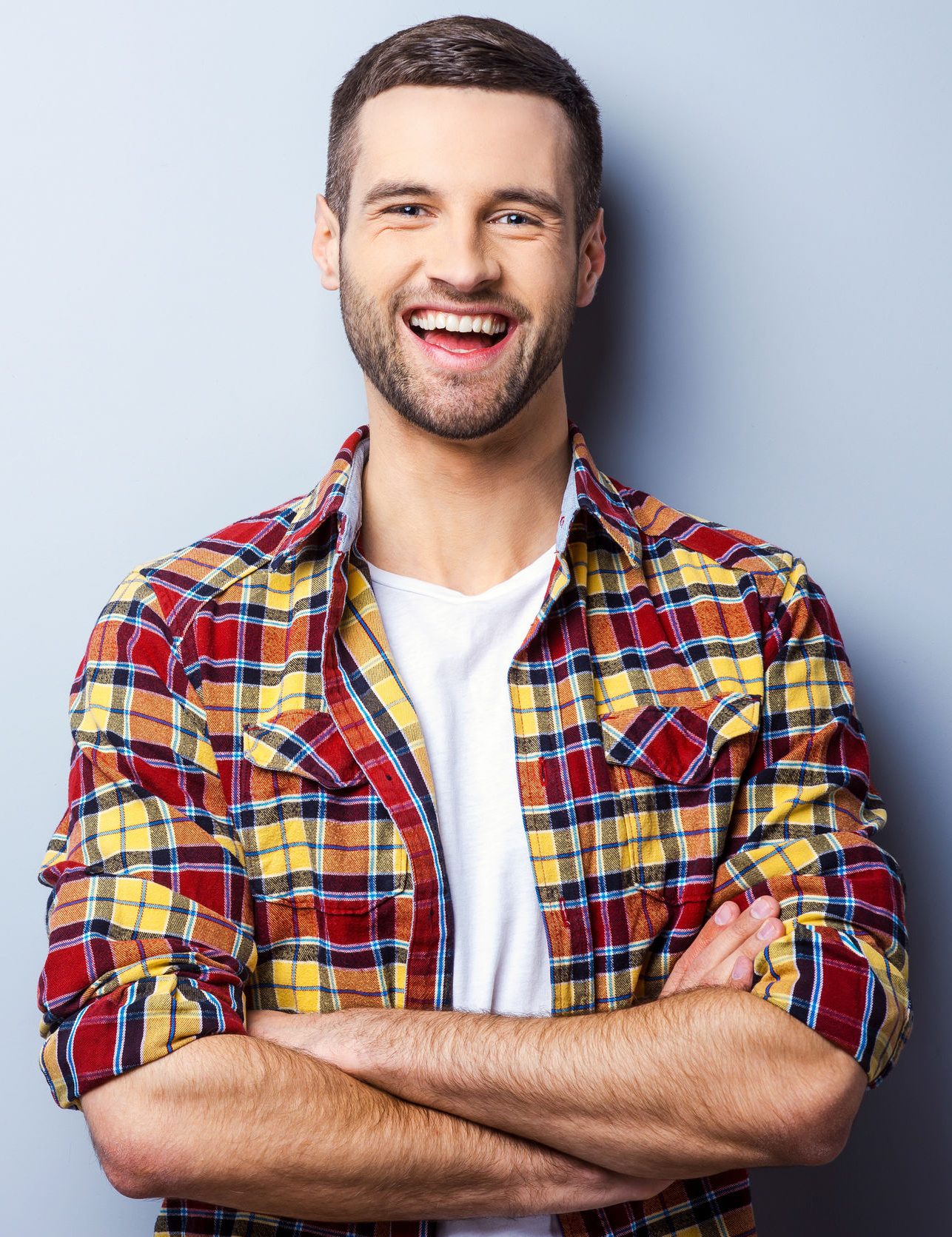 Happy young man. Portrait of handsome young man in casual shirt keeping arms crossed and smiling while standing against grey background